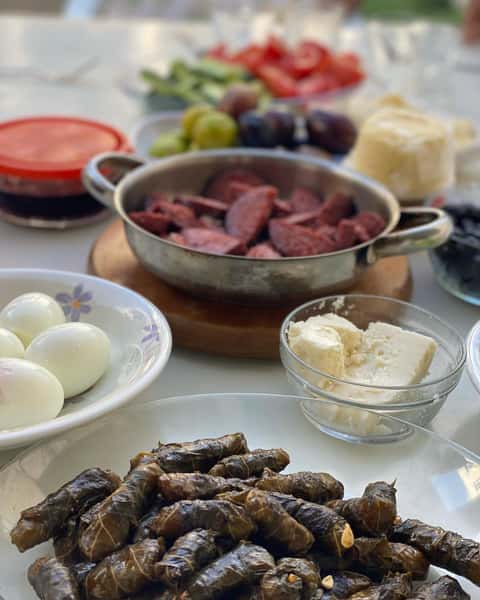 Traditional Turkish breakfast spread featuring homemade stuffed grape leaves (dolma), sliced Turkish sausage (sucuk), boiled eggs, feta cheese, olives, fresh vegetables, and breakfast accompaniments on a table at Ebby's Turkish Kitchen.