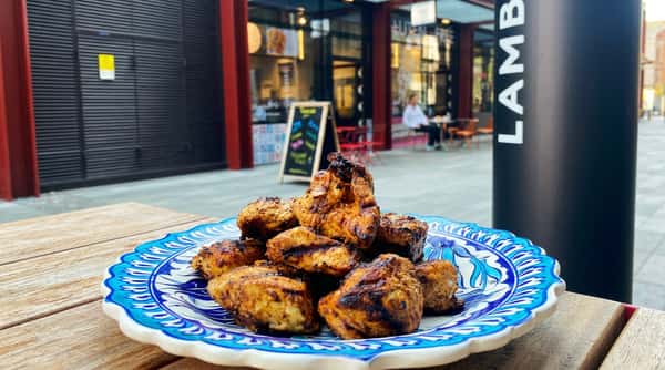 Grilled chicken pieces on a blue and white patterned plate, with a building and "LAMB STREET" sign in the background.