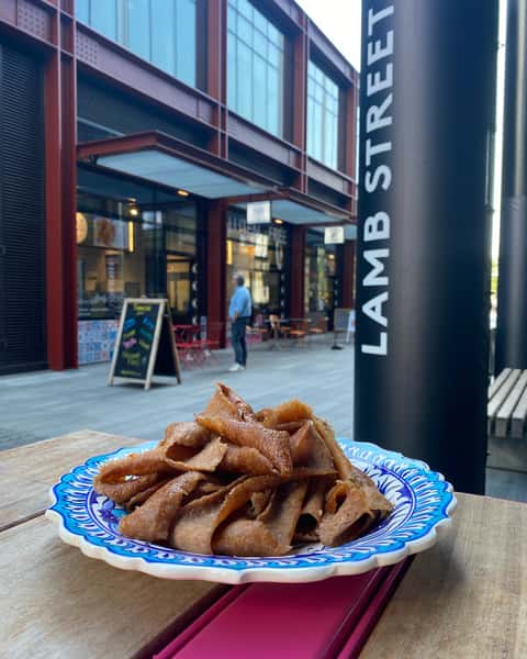 Shaved doner kebab meat on a blue and white decorative plate, set on a wooden table, with "LAMB STREET" sign and buildings in the background.