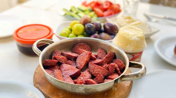 Sautéed Turkish sucuk (spicy sausage) slices in a pan, with olives and tomatoes in the background.