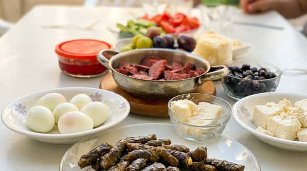 A table spread with various dishes including stuffed grape leaves, boiled eggs, cubed cheese, sliced sausage, and olives.