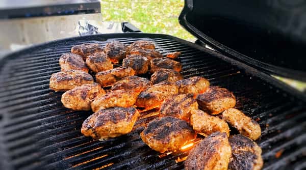 Browned meat patties grilling on an outdoor barbecue with flames visible through the grates.