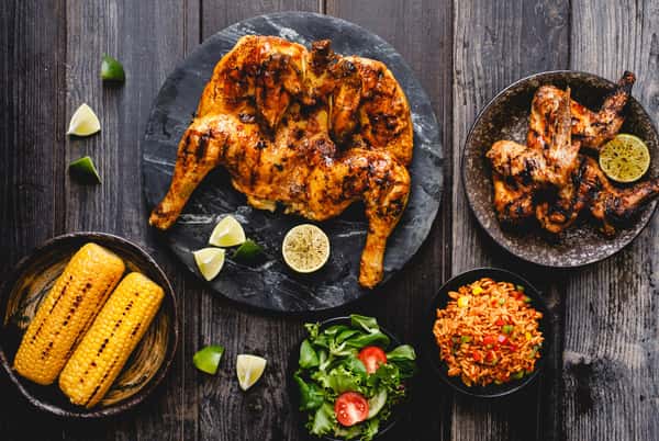 Overhead view of grilled spatchcock chicken with charred wings, lime wedges, corn on the cob, rice, and salad on rustic table