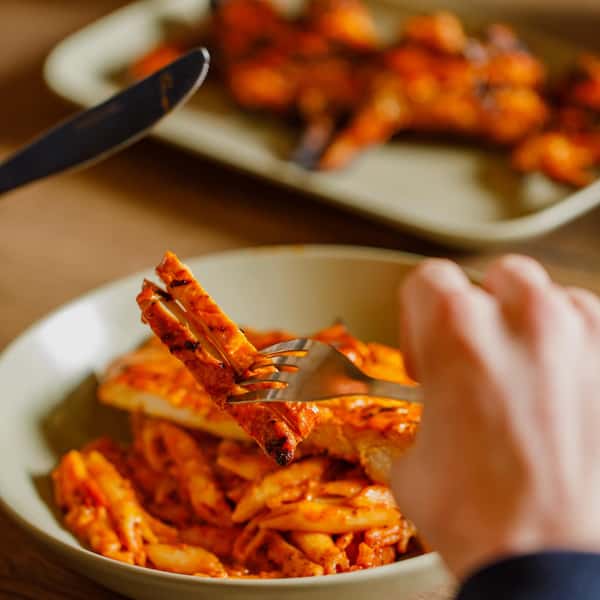 Fork lifting grilled chicken strips over a bowl of penne pasta in spicy tomato sauce, with plated chicken blurred in background