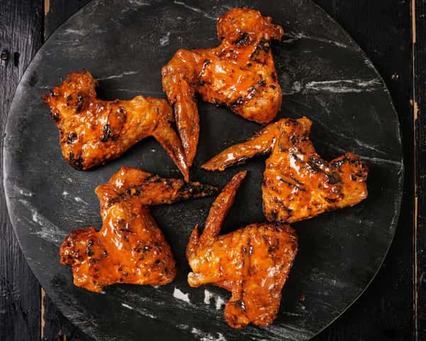 Six grilled chicken wings coated in glossy spicy orange sauce arranged on a dark round plate against a rustic black surface