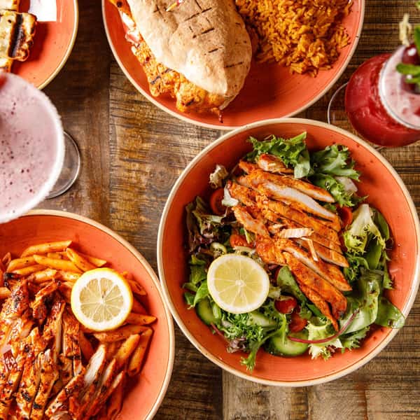 Overhead spread of grilled chicken salad with lemon, pita wrap and spiced rice, pasta, and pink cocktails on rustic table