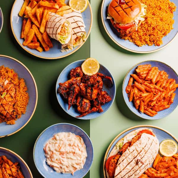 Overhead spread of grilled chicken wings, wraps, burgers, fries, rice, pasta, and sides arranged on blue plates over green background