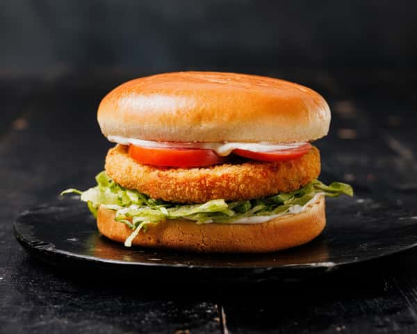 Breaded chicken sandwich with lettuce, tomato, and creamy sauce on a toasted bun, served on a dark plate.