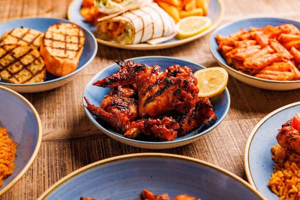 Assorted grilled chicken wings in bowls with lemon, fries, wraps, rice, and grilled vegetables on a wooden table.