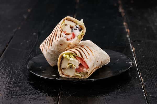 Two grilled chicken wraps with char marks, sliced to show lettuce, tomato, and creamy sauce on a black plate over a dark wooden table
