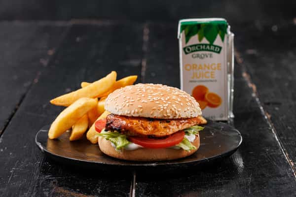 Grilled chicken burger with lettuce and tomato, served with thick-cut fries and a carton of orange juice on a dark wooden table