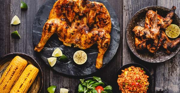 Overhead view of grilled spatchcock chicken with charred wings, lime wedges, corn on the cob, rice, and salad on rustic table