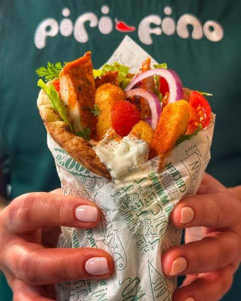 Hands holding a chicken wrap with seasoned fries, lettuce, tomato, red onion and creamy sauce, wrapped in Pico Fino branded paper