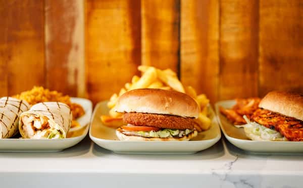 Three plated meals on a counter featuring a chicken wrap, crispy chicken burger, fries, and grilled chicken against a wooden backdrop