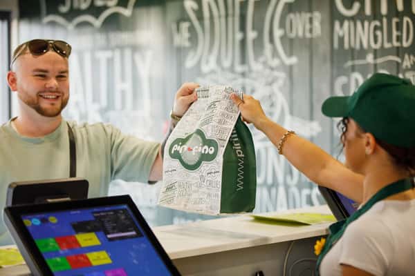 Smiling customer receives a branded takeaway bag from a staff member at a counter inside a casual restaurant setting