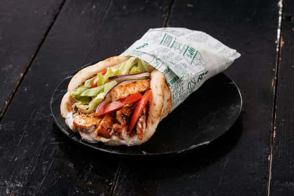 Chicken shawarma wrap filled with grilled chicken, lettuce, tomato, onion, and sauce, wrapped in printed paper on a dark wooden table.