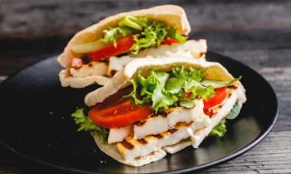 Pita sandwich filled with grilled cheese, fresh lettuce, and tomato slices, served on a black plate against a rustic wooden background