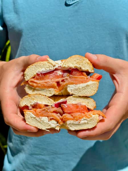 Hand holding a stacked bagel sandwich filled with smoked salmon, tomato, red onion, and cream cheese, captured against a sunny outdoor backdrop.