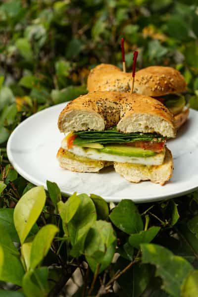 Veggie breakfast bagel sandwich on a white plate, filled with avocado, tomato, spinach, and egg, served outdoors against a leafy green background.