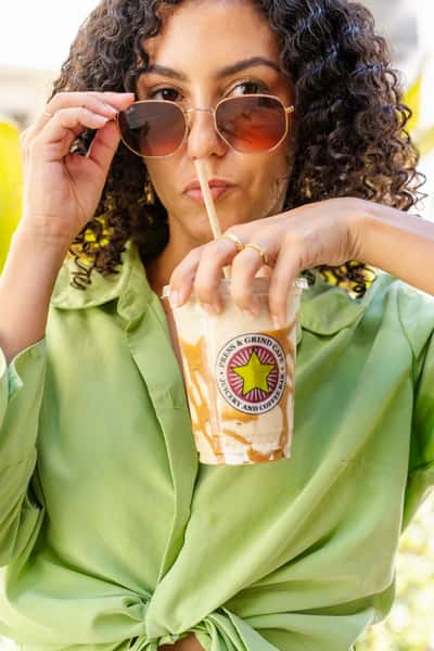Woman in a green blouse sipping a caramel-drizzled iced coffee from a Press & Grind Café cup, wearing stylish sunglasses in an outdoor setting.