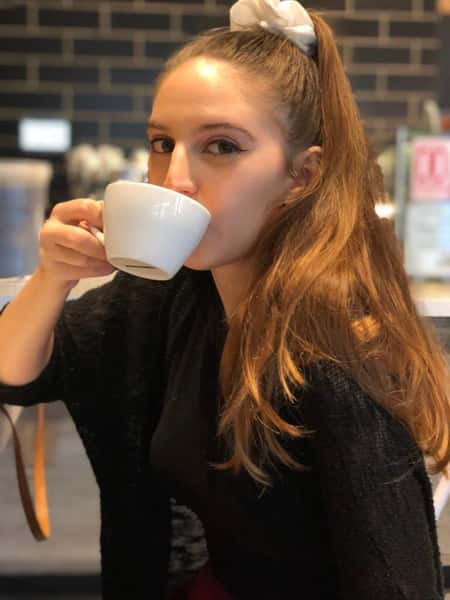 Woman sipping a hot coffee from a white ceramic cup in a cozy café, with a warm ambiance and modern tile backdrop.