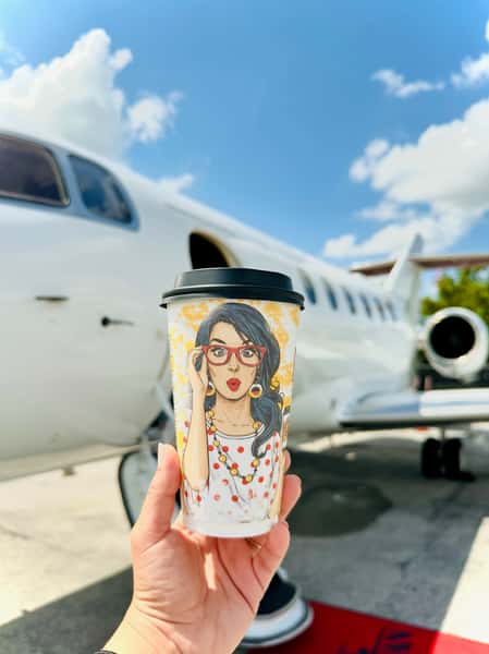 Hand holding a takeaway coffee cup with illustrated artwork in front of a sleek private jet on a sunny day, blending luxury and café culture.