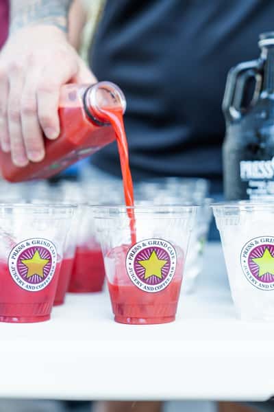 Bright red cold-pressed juice being poured into branded plastic cups at an outdoor café event, with a black growler jug in the background.