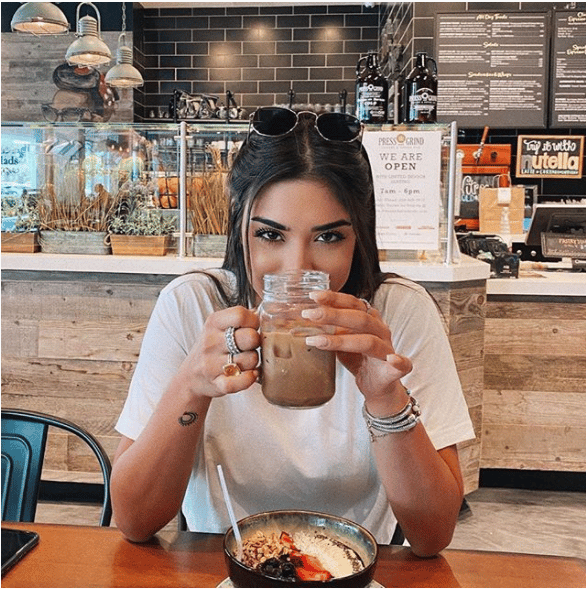 Woman enjoying iced coffee from a mason jar in a cozy café, seated in front of a granola-topped acai bowl, surrounded by rustic interior decor.