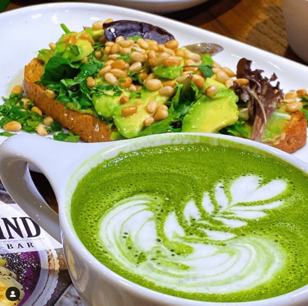 Artisan avocado toast topped with greens, pine nuts, and herbs, paired with a frothy matcha latte with latte art in a cozy café setting.