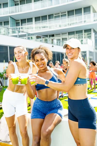 Three athletic women enjoying iced green juices on a rooftop during a sunny outdoor fitness event, smiling in activewear with a modern city backdrop.