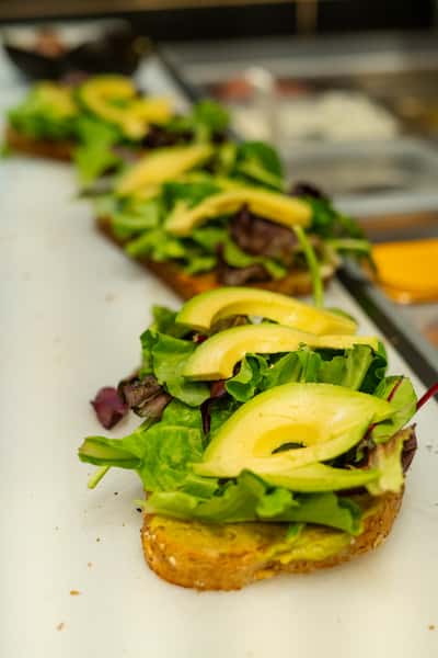 Fresh avocado toast being prepared in a café kitchen, topped with creamy avocado slices and mixed greens on whole grain bread.