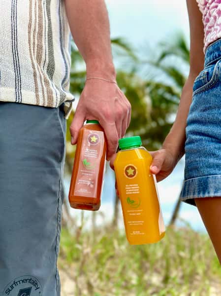 Two people holding cold-pressed juice bottles at the beach, featuring mango turmeric and green blends from a health-focused juice brand.