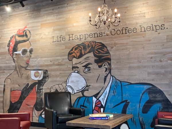 Artistic coffee shop wall mural featuring retro characters drinking coffee, with the quote "Life Happens... Coffee helps." and a chandelier overhead.