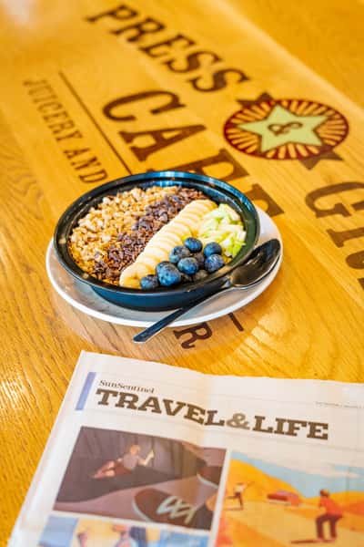 Colorful acai bowl topped with banana slices, granola, blueberries, and diced green apple, served on a wooden café table beside a travel newspaper.