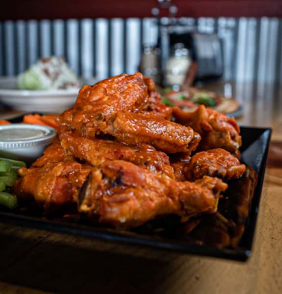 A close-up of a stack of crispy glazed chicken wings served on a black tray. The wings are coated in a shiny, flavorful sauce and garnished with chopped herbs, with small dipping sauce cups and a blurred restaurant background adding to the warm, casual dining atmosphere.