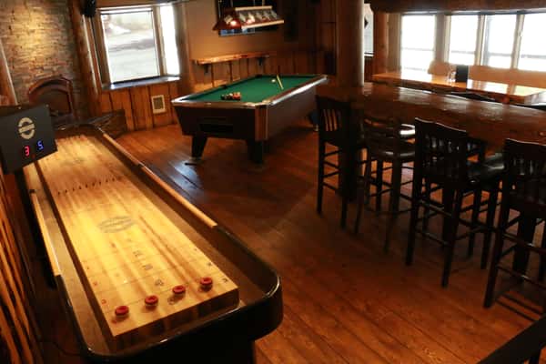A cozy game room inside a bar or pub featuring a green felt pool table and a wooden shuffleboard table. Warm wood flooring, high-top seating, and natural light from the windows create a relaxed and inviting atmosphere perfect for casual games and social gatherings.