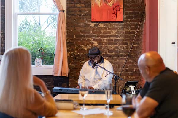 A musician in a white shirt and fedora plays the keyboard and sings into a microphone, performing live for guests at an intimate venue with exposed brick walls and soft lighting.