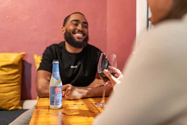 Smiling man at a table with a Delirium Tremens beer bottle and a woman holding a glass of red wine in the foreground.