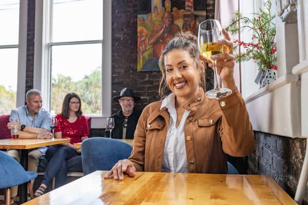 Smiling woman in a tan jacket raises a glass of white wine in a cheerful toast, with friends chatting in the background.