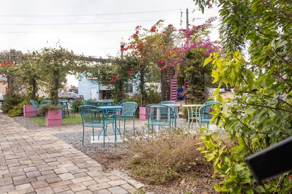 Outdoor dining area with teal chairs, pastel tables, brick walkway, and vibrant flowering vines under wooden pergolas.