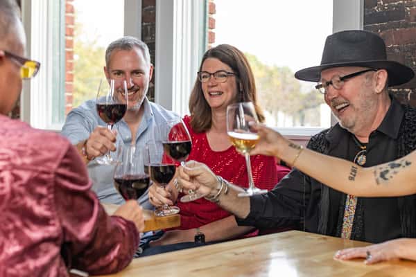 Group of friends cheerfully toasting with red and white wine near sunlit windows in a cozy restaurant.