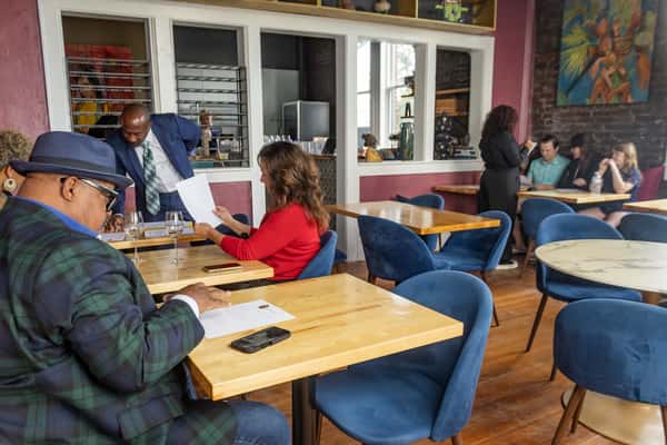 Guests in a stylish restaurant read menus and chat as servers take orders; blue velvet chairs and vibrant artwork decorate the space.