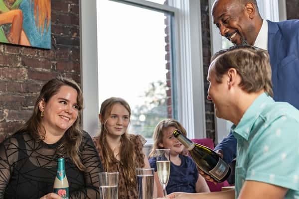Smiling server pours sparkling wine for a cheerful family seated by large windows in a cozy brick-walled restaurant.