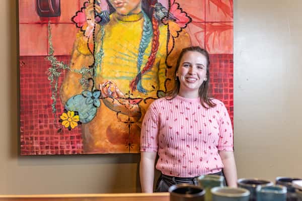 Smiling woman in a pink sweater stands in front of a colorful painting with braided hair and floral accents.