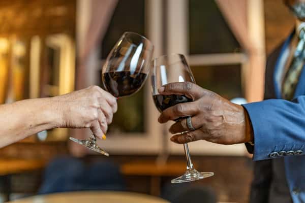 Close-up of two people clinking glasses of red wine, one in a blue suit and the other with yellow-painted nails, in a cozy indoor setting.