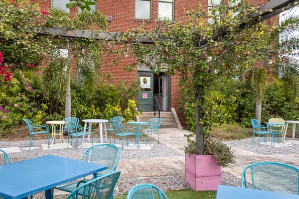Outdoor patio with colorful metal chairs and tables, lush greenery, and a view of the entrance to Delicious restaurant.