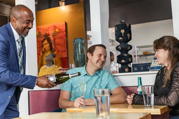 Waiter pours sparkling wine for a smiling couple seated at a cozy table in a stylish restaurant with eclectic decor.