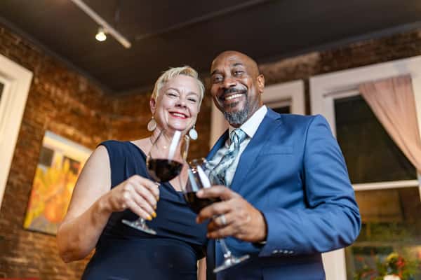 Smiling couple dressed elegantly clinking glasses of red wine in a warmly lit room with brick walls.