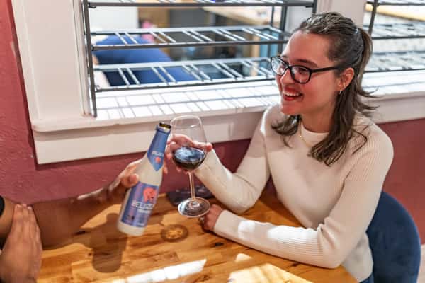 Smiling woman with glasses holds a glass of red wine while chatting with someone offering a Delirium Tremens beer.