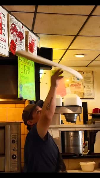 Pizza maker tossing a large flat pizza dough in the air within a restaurant kitchen.